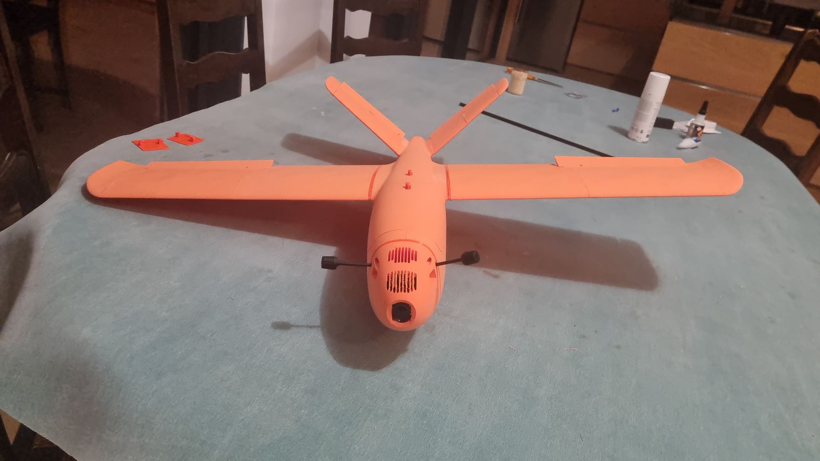 The assembled orange plane on a light blue-grey tablecloth, seen from the front. The wings and the V-shaped tail are attached. The nose has an open camera hole with vents above it, and a black rod sticks out from each side of the nose. A few spare orange printed bits, a carbon rod and some glue bottles lie on the table.