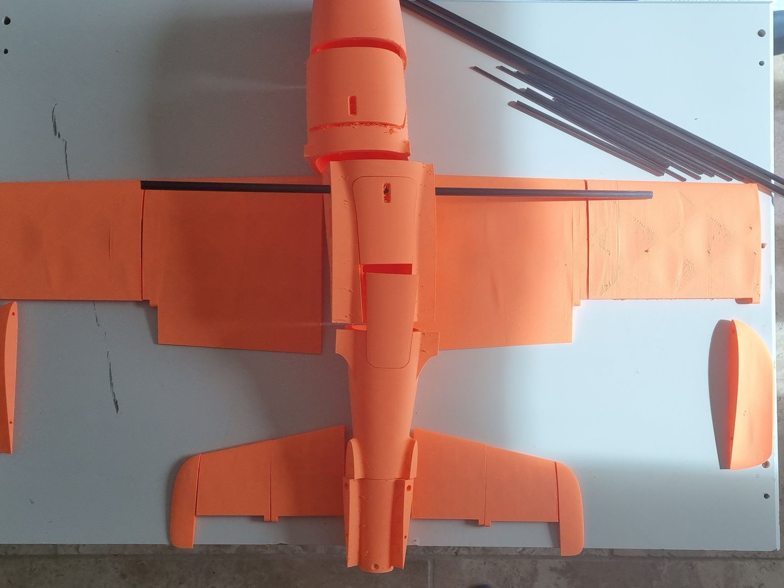 Top view of the printed parts laid out on a white board in the shape of the plane: fuselage sections down the middle, wing panels left and right with a dark carbon rod through them, the tail pieces at the bottom, and a bundle of thin carbon rods in the top right corner.