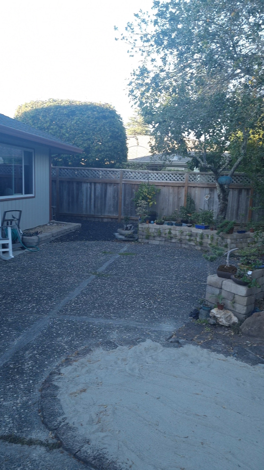 A gravel patio with a sandy circle, a wooden fence and the corner of a house, in warm evening light.