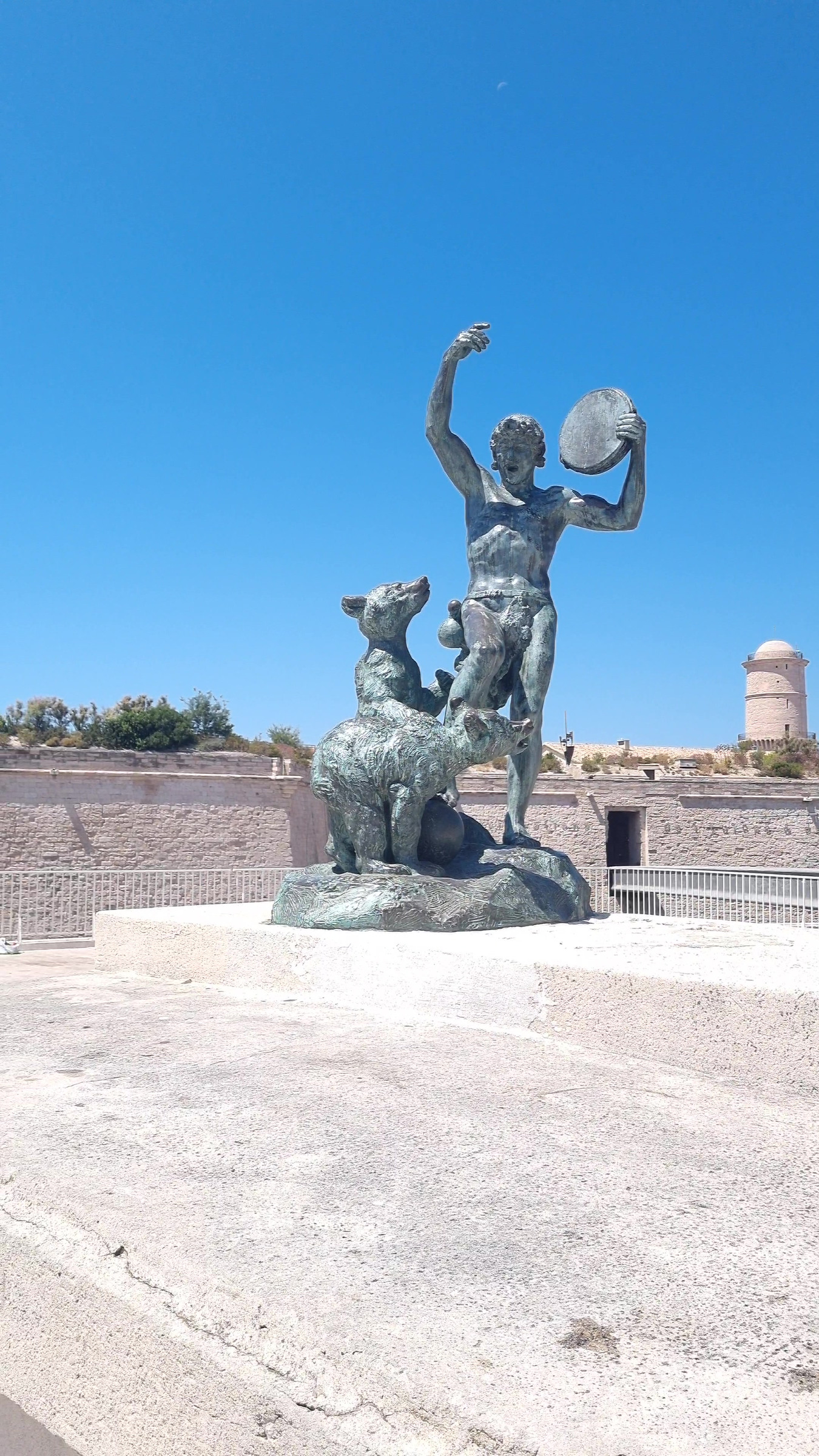A bronze statue of a man raising a tambourine, with two bears at his feet, on a stone terrace under a blue sky.